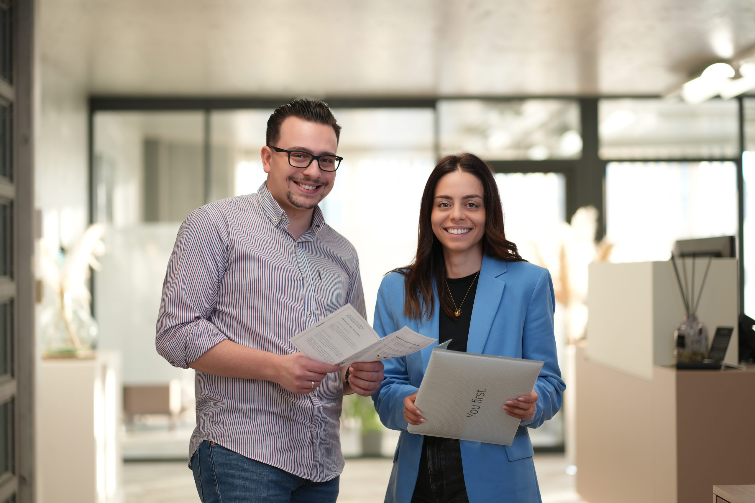 Joel Cardoso et Katia Pinto de notre agence d'Yverdon-les-Bains qui pose dans le hall de la réception. Ils regardent la camera et ont un flyer te une enveloppe à bulles New Work dans les mains